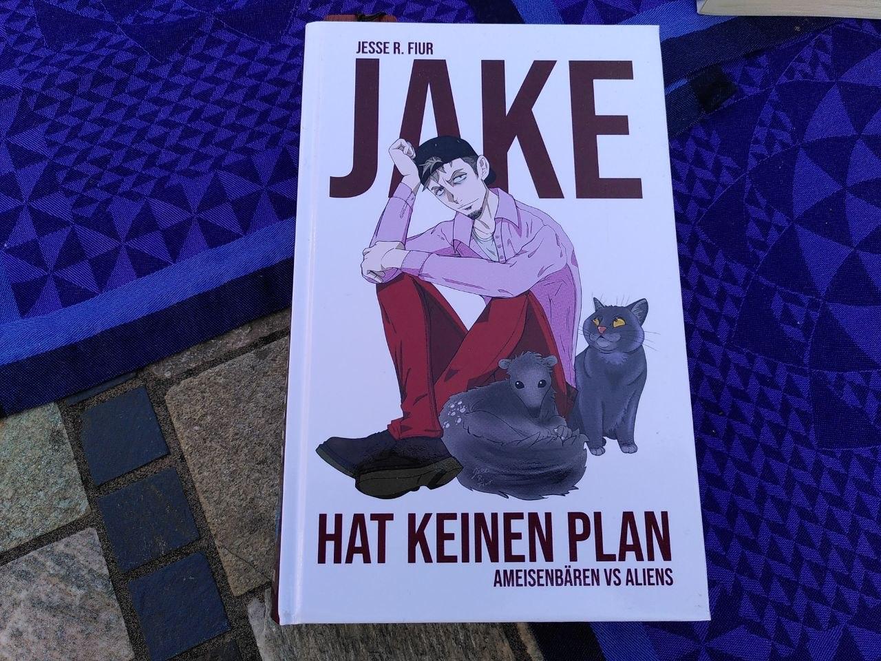 Das Buch »Jake hat keinen Plan – Ameisenbären vs Aliens« von Jesse R. Fiur liegt als Hardcover auf einem mit Tischsets arrangierten Fliesentisch.